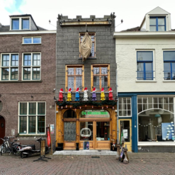 Foto van de gevel van de winkel genaamd De Stoelendans en Masterpieces gelegen aan de Nieuwstraat in Deventer. De afbeelding toont een houten gevel met daarop zeven gekleurde kabouters. Voor de deur staan twee gekleurde vlaggen, drie houten paddenstoelen en twee stoepborden. Aan de gevel hangt een grote jutte zak voor Sinterklaas. De kleur van de gevel is bruin met grijs. Het gebouw heeft twee deuren en vier ramen.